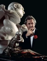 Franco Maria Ricci fotografato col celebre fiore di bachelite all’occhiello e alcune opere novecentesche della sua collezione, il Vir Temporis Acti di Wildt e una statuetta Déco